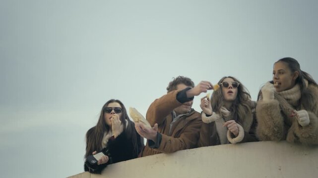 A group of young friends enjoys sharing snacks and laughter outdoors. They are seen bonding and having a good time together in a casual setting.