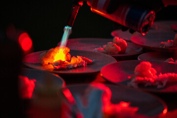 A chef uses a culinary torch to flambé a dish on a dark plate, creating a dramatic flame. Several...