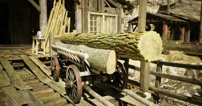 A wooden cart transports large logs at a rustic sawmill surrounded by wooden structures and rocky terrain. The vibrant sky adds to the historic ambiance of the setting.