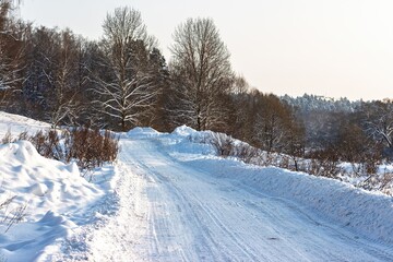 Obraz premium Evening sun bathes a winding snowy road, tire tracks tracing through fresh powder. Winter landscape offers serene solitude amidst bare trees and frosty air