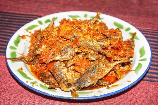 Close-up of traditional Indonesian food, ikan balado or small fried fish with spicy red chili sauce served on a white enamel plate with green edges, on a red striped tablecloth.