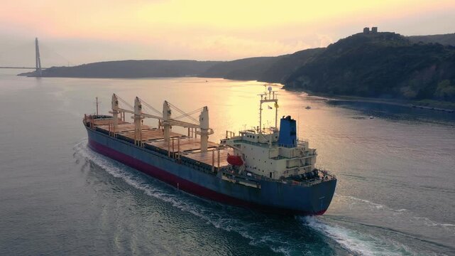 Bulk carrier with deck cranes navigating strait toward suspension bridge at golden sunset with hilltop fortress in background. Aerial view