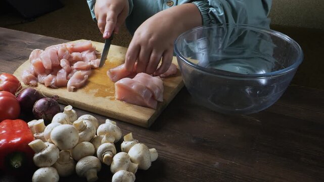 A woman's hands slice chicken fillet on a cutting board on a table. Medium shot.