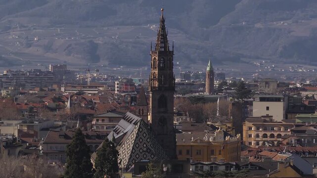 Aerial pan and zoom over Bolzano, Italy, with Gothic spire, diamond roof, terracotta homes, neoclassical facade, brick tower, and Alpine ridges in late day light.