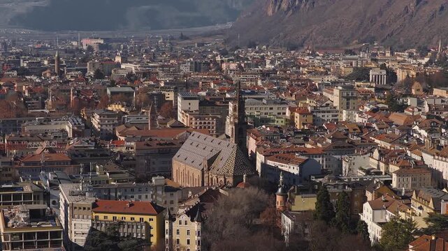 Aerial pan shows Bolzano, Italy, the Gothic cathedral with diamond roof and spire, red tile roofs, narrow streets, plazas, and Alpine foothills in warm late afternoon light.