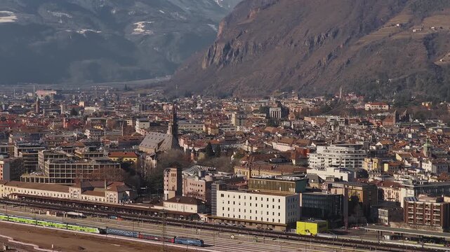 Aerial sweep over Bolzano, South Tyrol, shows Hauptbahnhof, green regional trains, a colorful locomotive, and the cathedral spire beneath Alpine slopes and winter light.