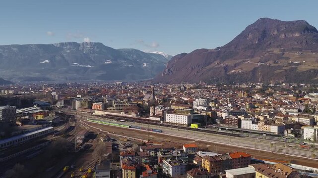 Aerial winter panorama of Bolzano, Italy, with the central railway station, active rail yard, green regional trains, historic town center, and terraced Alpine slopes.