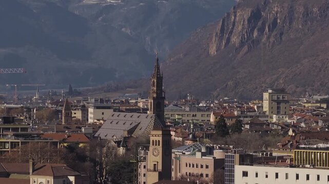 Aerial view of Bolzano, Italy, with Duomo spire and diamond roof. Camera pans to alpine cliffs and Adige valley. Warm low light, wide frame, bare trees, cranes.