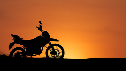 Silhouette of motorcycle set against a vibrant sunset sky. The motorcycle stands still on a sandy terrain © Stock4u2024 