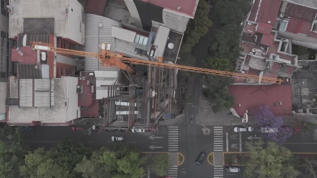 Top-down aerial view of a construction crane over an urban building site, showing the structure under development, surrounding streets, and city infrastructure.