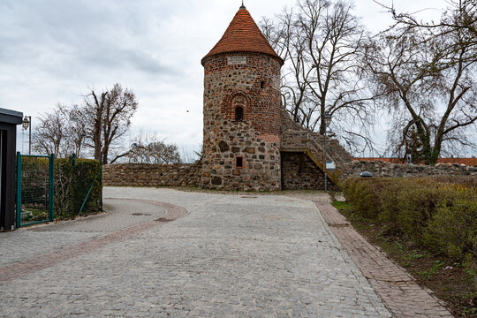 A gently curved cobblestone street leads toward a historic round tower that once formed part of the town&rsquo;s medieval fortifications. The fieldstone masonry and tiled roof reflect traditional defensive 