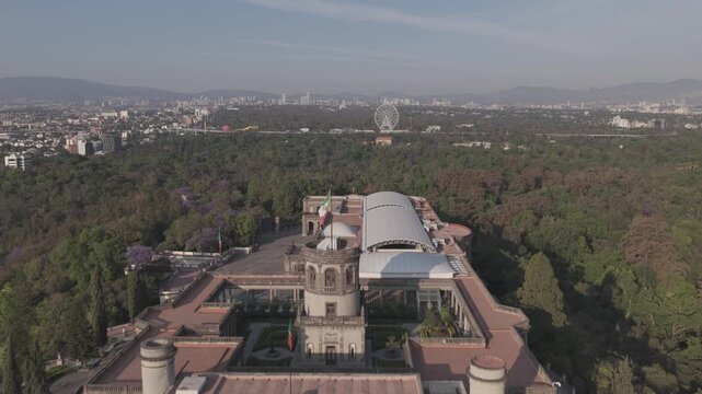 Aerial view over Chapultepec Castle at sunrise with a panoramic view of Bosque de Chapultepec and the Aztl&aacute;n Ferris wheel visible in the distance under a clear blue sky.