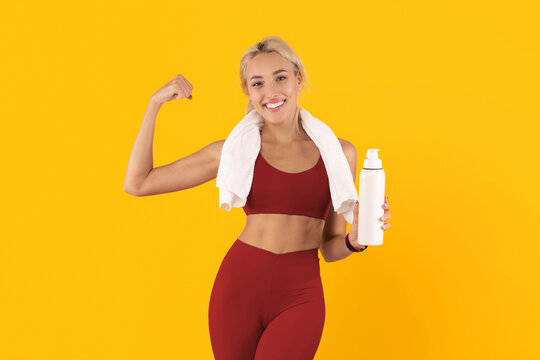 A woman smiles and flexes her arm while holding a water bottle in her other hand. She wears workout clothes and has a towel draped around her neck. The background is bright yellow.