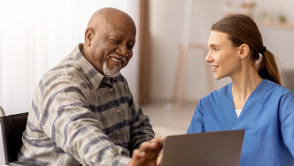 A caregiver and an older man are sitting together in a room. The man smiles as the caregiver shows him something on her laptop. They are engaged in conversation and share a moment of connection.