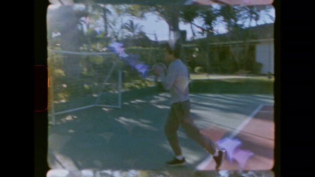 Authentic vintage 8mm film footage of a young man dribbling a basketball on a residential court. He moves across the court on a sunny day. The camera tilts up to the hoop. Nostalgic and retro feel.