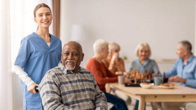 A caregiver assists an elderly man in a senior care center. Other residents play chess and socialize at tables. The space is bright with large windows. Various activities are happening.