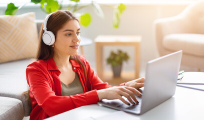 A young woman sits on the floor in a cozy area, engaging with her laptop. She wears headphones and...