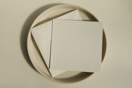 Summer minimal wedding stationery. Blank square greeting card mockup with beige envelope, invitation. Ceramic dessert plate in soft sunlight. Table background with shadows overlay. Flatlay, top
