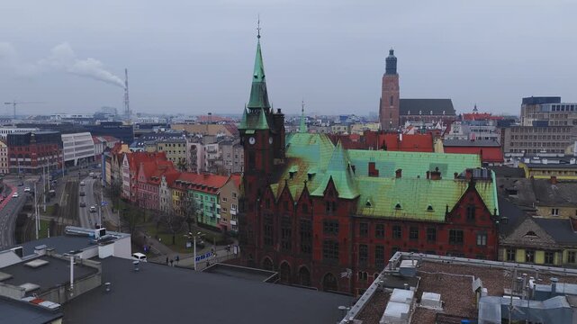 Aerial view of central Wroclaw, Poland shows red brick clock tower, gabled townhouses, curving tram tracks, moving cars and trams, and a church under overcast skies.