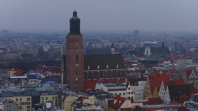 Aerial pan over Wroclaw, Poland, shows a tall brick Gothic church, red tiled gabled roofs, dense historic blocks, and a modern skyline fading into gray haze.