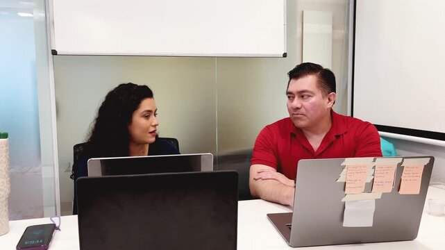 Smiling woman and man two professionals discussing ideas while working on computers at office desk in Brazil look down camera medium shot