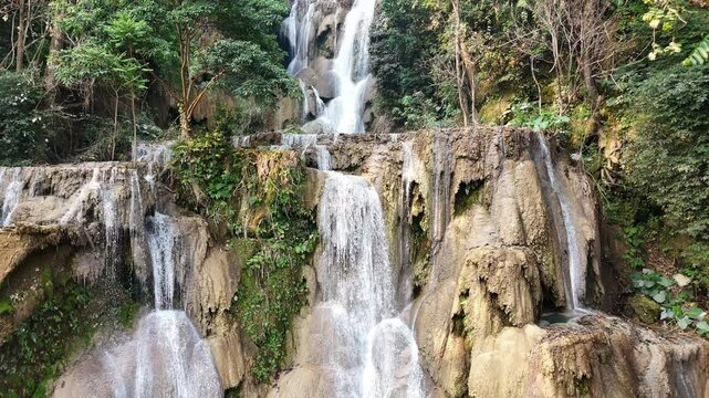 Aerial drone view of Kuang Si Waterfall main cascade near Luang Prabang, where multiple streams of water fall down limestone rock formations into turquoise pools surrounded by dense jungle