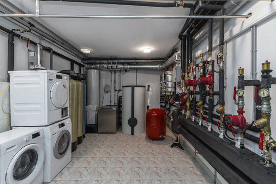 Laundry machines are placed in a basement area along with water tanks and pipes. The space is functional with tools for managing water and energy needs in the household