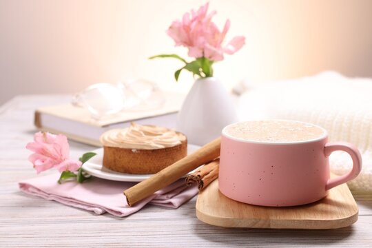 Cup of coffee, tasty tart, cinnamon sticks and vase with flowers on light wooden table, closeup