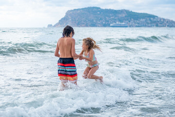 Two young children boy and girl, holding hands and playing in ocean waves together, siblings enjoy...