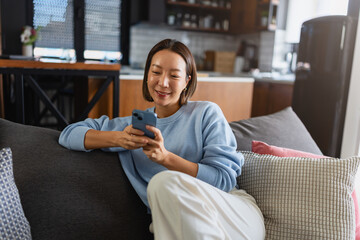 Young East Asian woman relaxing on a sofa, smiling while using her smartphone. Cozy home setting,...