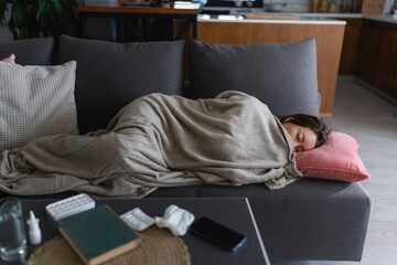 Young East Asian woman sleeping on sofa under cozy blanket, resting peacefully at home after a long...