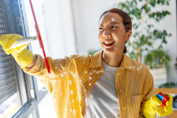 Smiling Asian woman cleaning window with spray and cloth in bright sunlight. Cheerful home cleaning...