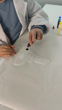 A young student wearing a white laboratory coat carefully drips red liquid colorant onto a smooth white slime base during a science experiment.