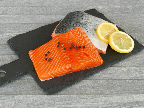 Two raw salmon fillets on a black slate platter with peppercorns and fresh lemon slices.