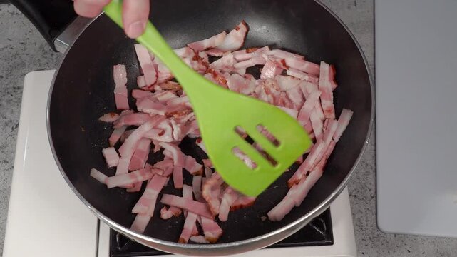 A deep frying pan of cut up bacon stirred by a green spatula with sizzle audio