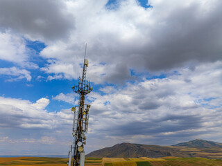 Obraz premium Telecommunications tower stands tall in expansive rural landscape. Multiple antennas and dishes are visible against a backdrop of cultivated fields and hills under a cloudy sky.