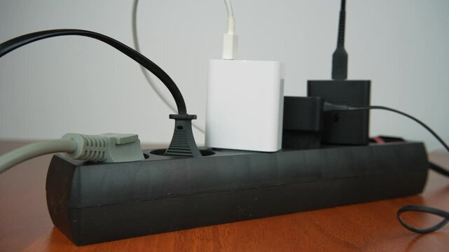 Close-up of a human hand plugging a power adapter into a black extension cord with multiple chargers connected. Concept of electricity usage, home office setup and energy consumption