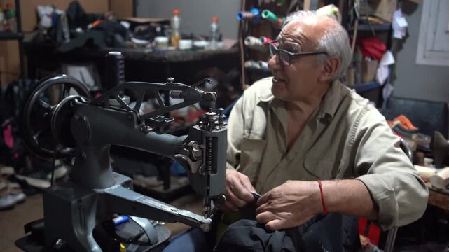 Older man cobbler sitting at sewing machine repairing leather backpack in home workshop, close-up of hands, thread, tools, and detailed artisan craftsmanship in action