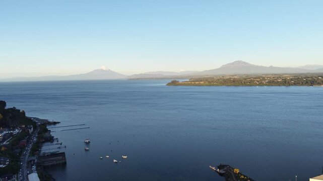 Cinematic aerial pulling back from the calm blue waters of Lake Llanquihue to reveal the city of Puerto Varas, featuring the snow-capped Osorno and Calbuco volcanoes in the background