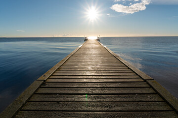 Obraz premium Straight wooden jetty extending toward the open sea with bright sun above the horizon. The calm water reflects the sky creating a peaceful marine landscape often associated with summer travel, relaxat