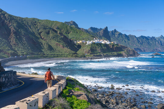 A tourist is hiking along a winding road by the ocean in Anaga Rural Park. In the distance &ndash; the village of Almaciga. Tenerife. Canary Islands.