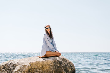 Woman Beach Smiling: Young woman in sunglasses sits on a large rock by the sparkling sea, enjoying a bright summer day. © svetograph