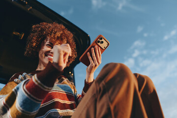 Happy woman with curly hair wearing rainbow sweater smiles holding smartphone near car outdoors...