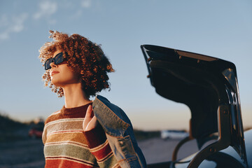 Woman with curly hair wearing sunglasses and a rainbow sweater stands near an open car trunk at sunset. The woman looks confident and relaxed in a casual lifestyle scene. © SHOTPRIME STUDIO