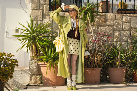 Stylish woman in green trench coat and retro outfit posing outdoors near stone wall