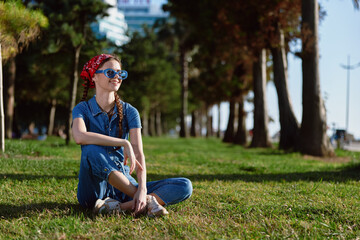 Lifestyle woman in modern boho-western denim street style sitting on grass outdoors, wearing sunglasses with film color effect, relaxed and smiling in sunny park environment.