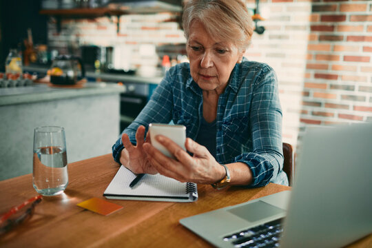 Senior woman managing finances on smartphone at kitchen table