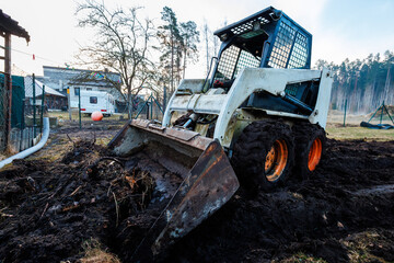 Fototapeta premium Skid Steer Loader Digs Soil in Rural Homestead with Trailer and Fence
