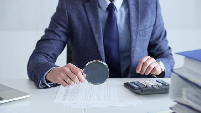 Businessman or accountant in a blue suit and tie sitting at a desk, using a magnifying glass to analyze financial documents with a calculator nearby, close up. Audit and taxes theme in business