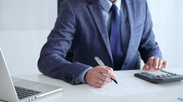 Businessman wearing suit and tie is using calculator and taking notes while working at desk in office, with laptop and stack of folders nearby, close up. Audit and taxes theme in business